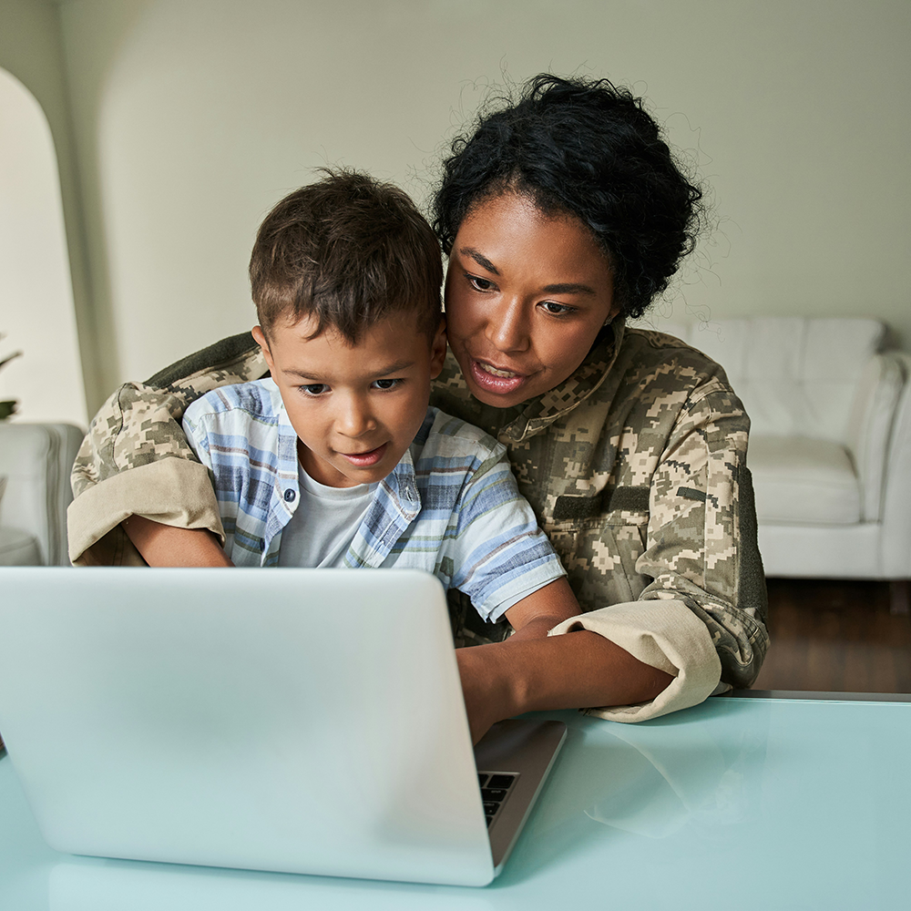 A person in military uniform uses a laptop with a young boy sitting on their lap, both focused on the screen.